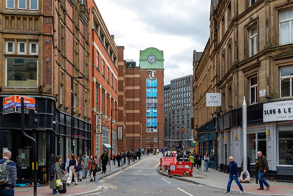 Urban street scene with brick buildings and pedestrians, featuring a clock tower in the distance on a cloudy day in Leeds, UK.