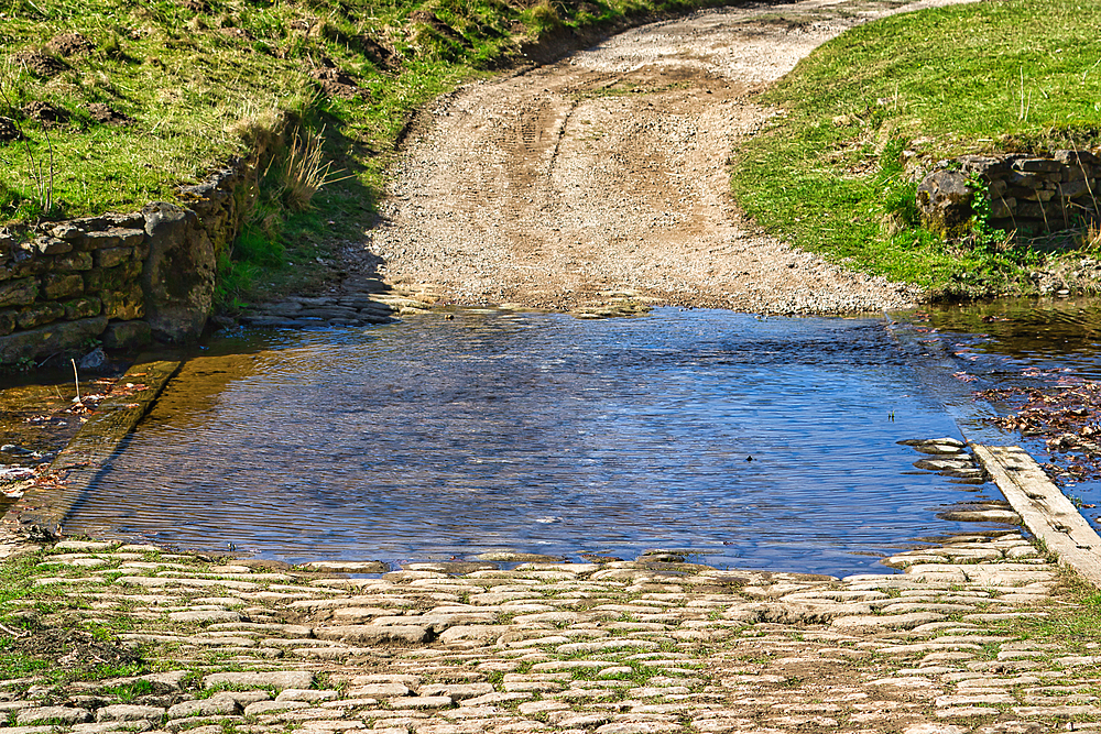 Low-angle view of a stream crossing a cobblestone road with a dirt road leading to it and grassy banks on either side at Studley Royal Park, UK.