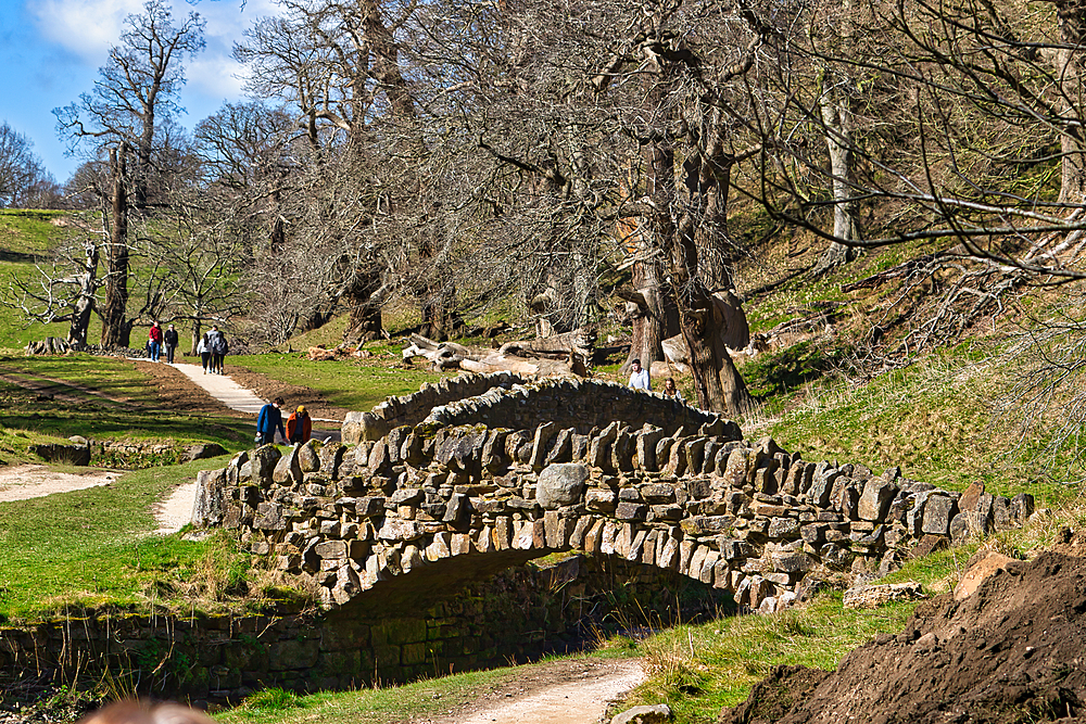 An old stone bridge over a dirt path in a park-like setting with leafless trees and people walking in the background at Studley Royal Park, UK.