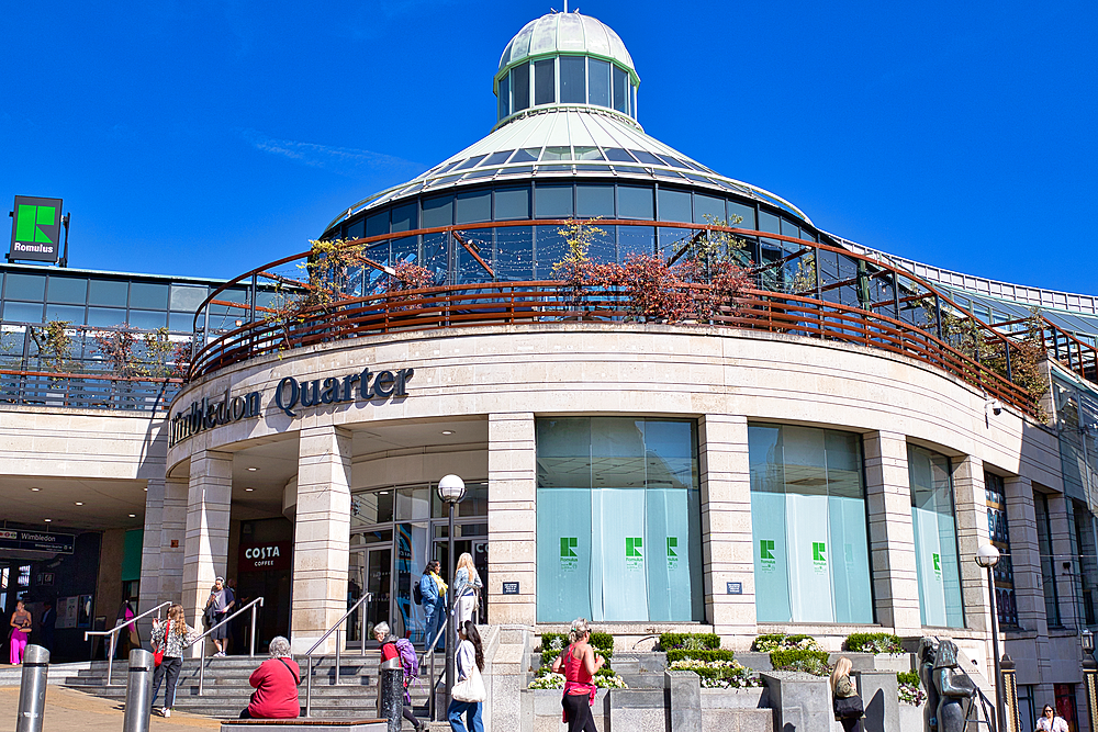 Exterior view of Wimbledon Quarter shopping centre on a sunny day. People are entering and exiting the building. Clear blue sky overhead.