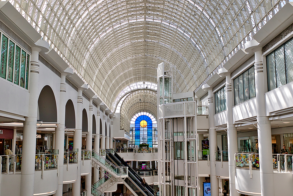Interior view of a multi-story shopping mall with a vaulted glass ceiling, columns, escalators, and a glass elevator shaft at Bentall Centre in Kingston, UK.