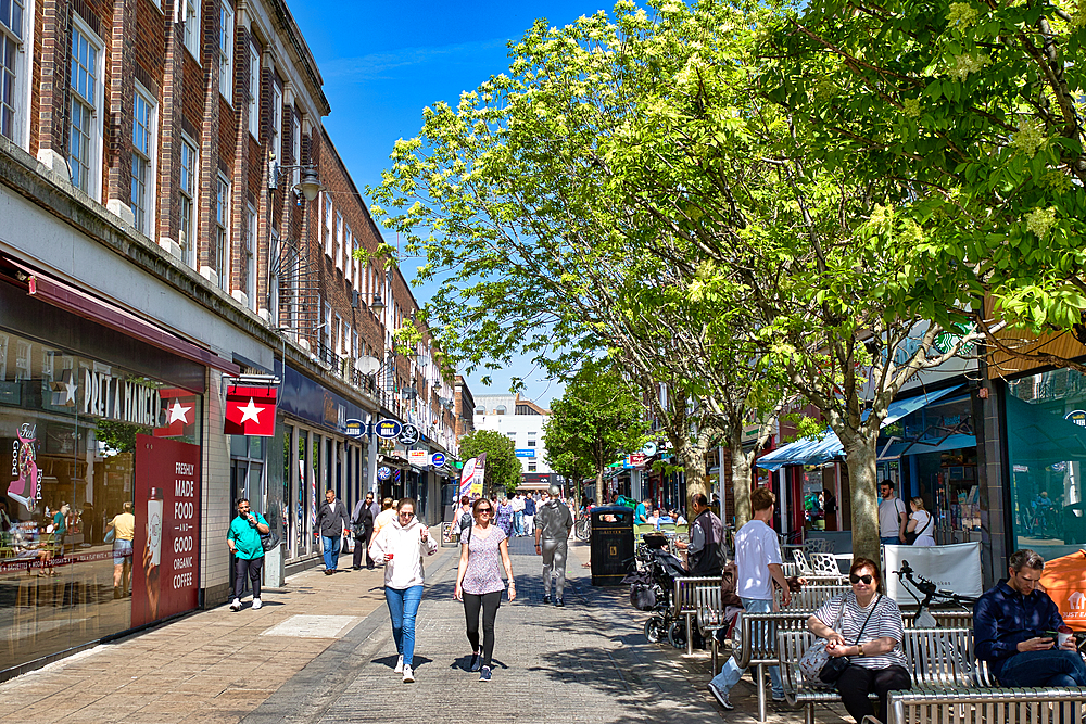 A bustling pedestrian street with shops, trees, and people walking and sitting under a clear blue sky in Kingston, UK.