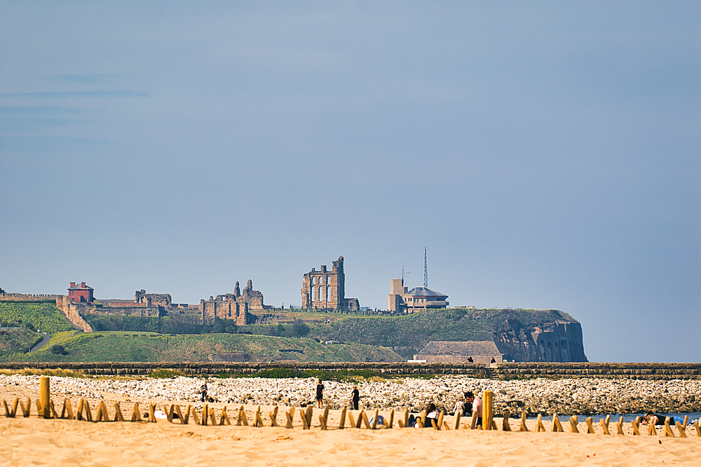 Coastal view of castle ruins atop a hill, people on a beach, and a protective barrier in the foreground at Sandhaven Beach, UK