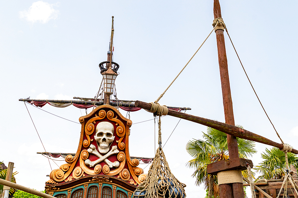 The image showcases the stern of a pirate ship with a skull and crossbones emblem, rigging, and mast against a pale blue sky at Disneyland Paris.