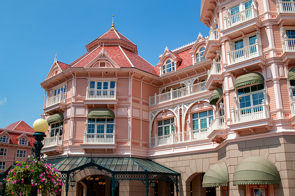 Facade of a pink building with white trim, red tile roof, green awnings, and balconies under a clear blue sky at Disneyland Paris.