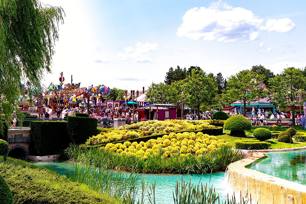 A vibrant park scene with water features, manicured hedges, a yellow flower bed, a carousel ride, and a crowd of people under a blue sky at Disneyland Paris.