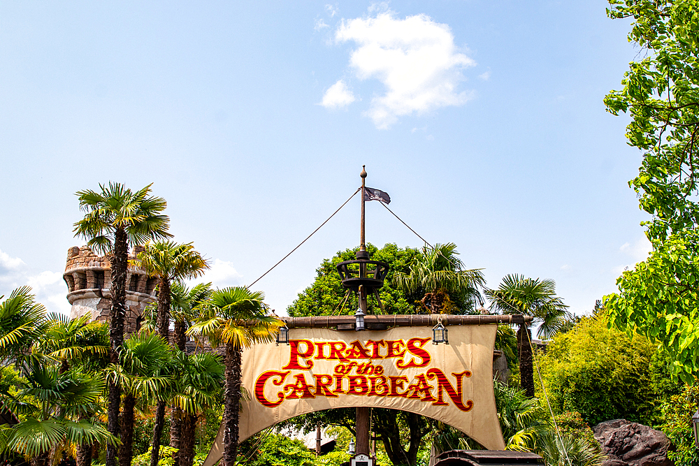 A shot showcasing the 'Pirates of the Caribbean' entrance sign, framed by palm trees, greenery, and a turret against a bright blue sky at Disneyland Paris.