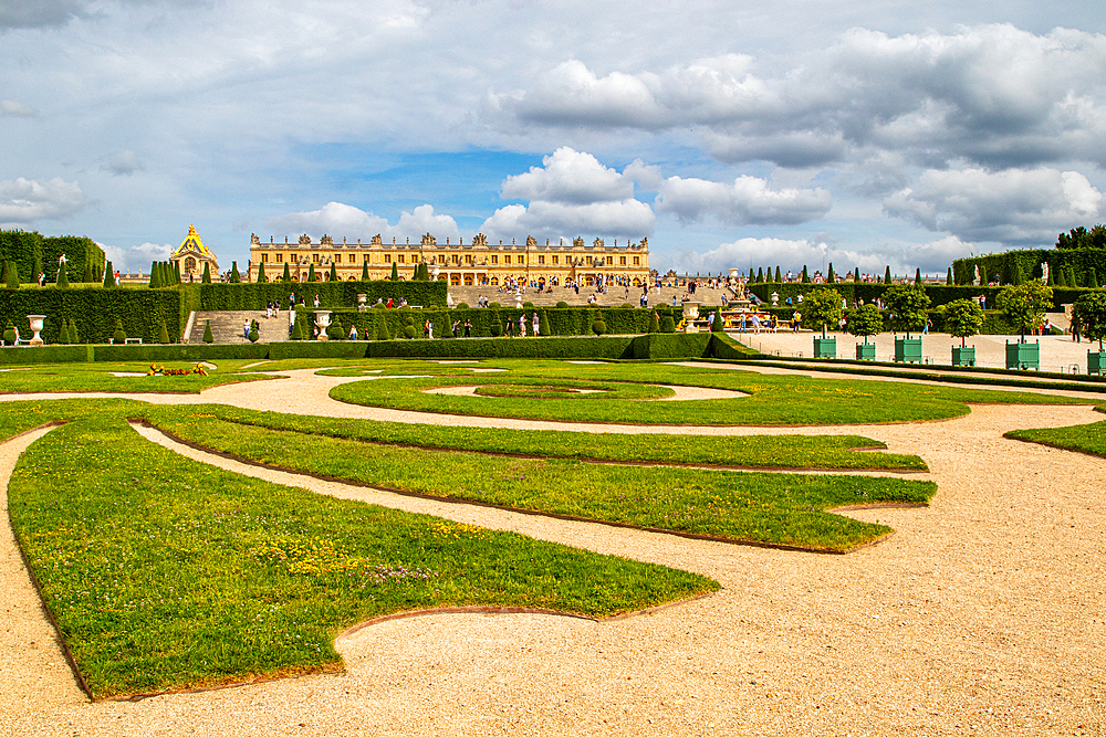 Versailles gardens with geometric lawns and gravel paths, leading to the palace and Orangerie under a partly cloudy sky at Palace of Versailles, Paris.