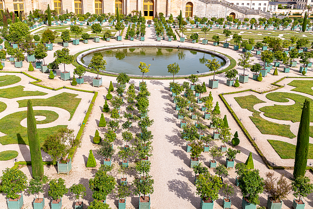 Aerial view of a formal garden with a circular pond, potted trees, topiary, and gravel paths, adjacent to a large building at Palace of Versailles, Paris.