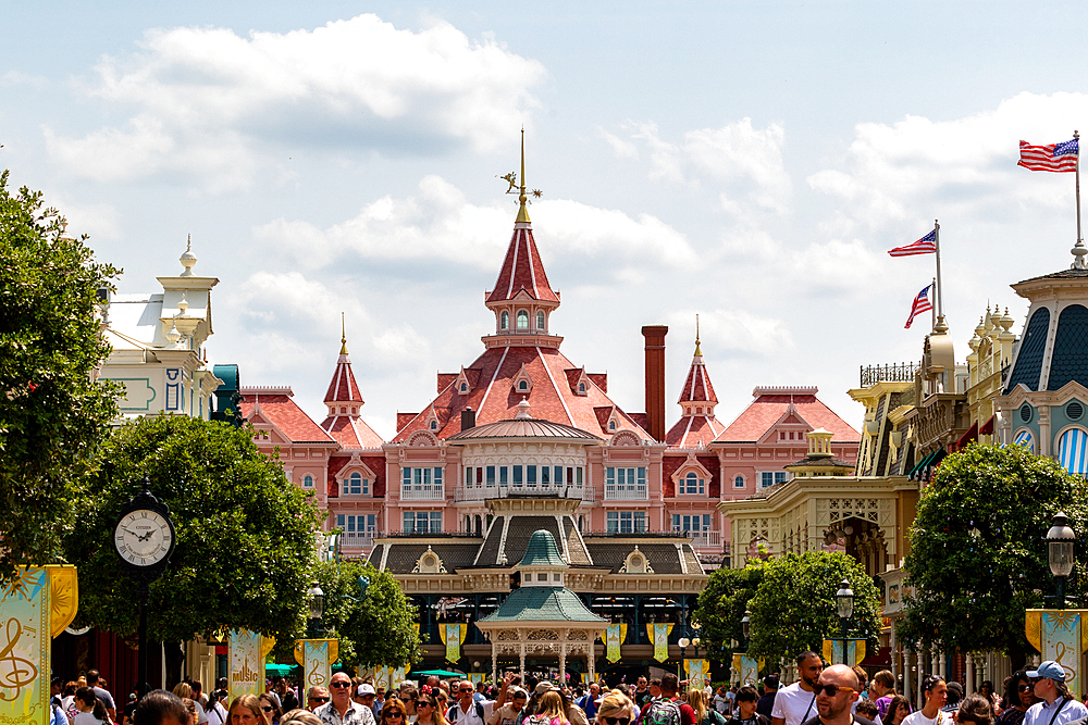 A crowd of people on a street lined with themed buildings, including a pink hotel with red roofs, under a partly cloudy sky at Disneyland Paris, France.