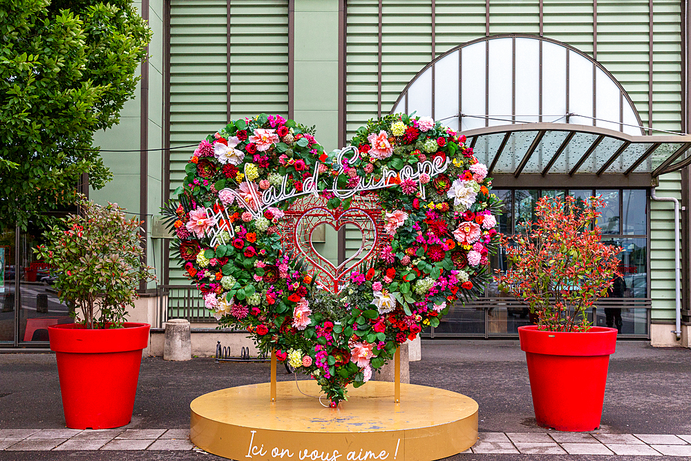 A large, heart-shaped floral display stands on a yellow platform in front of a building. Two large potted plants flank the display.