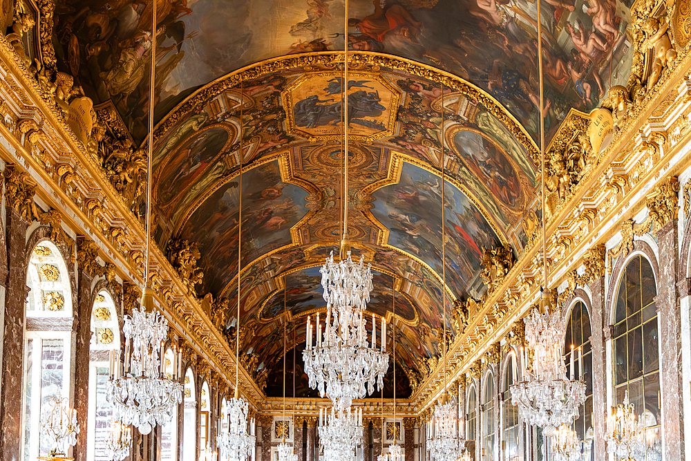 Ornate interior featuring a painted ceiling, chandeliers, and gilded details in a grand hall with arched windows and marble accents at Versailles Palace, Paris.