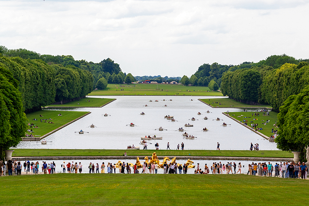 Wide angle shot of a large pond with rowboats, flanked by manicured grass and trees, viewed from a grassy hill with a crowd in front at Versailles Gardens, Paris.