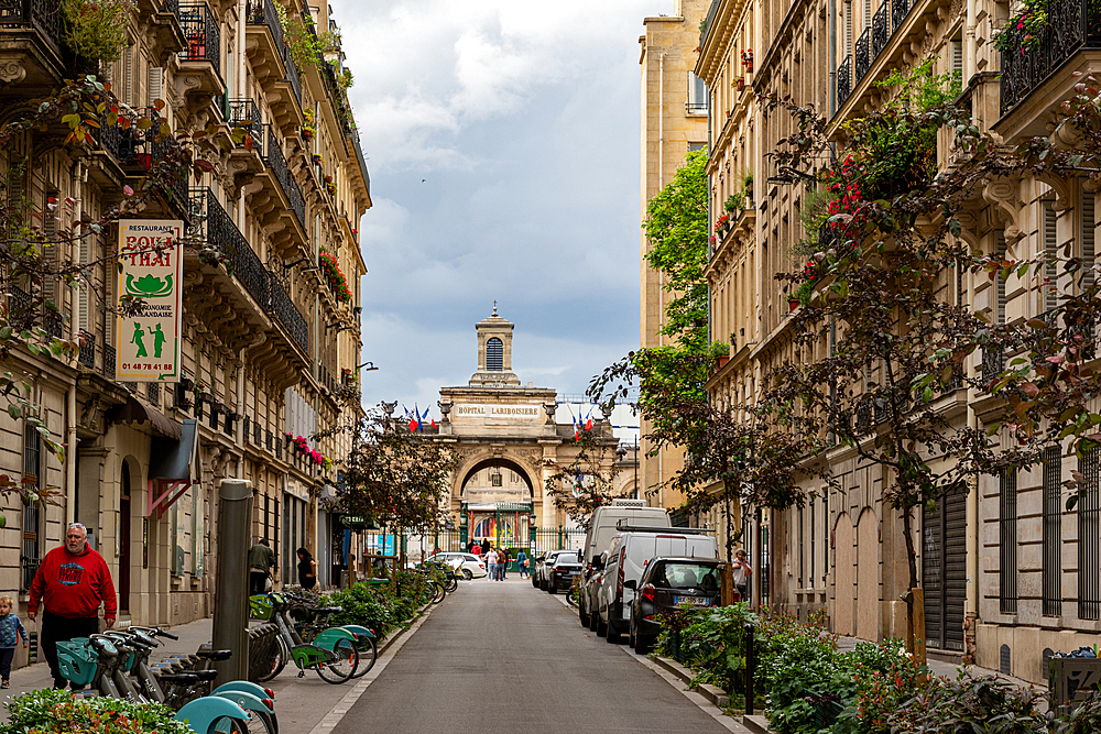 Parisian street view featuring architecture, parked vehicles, pedestrians, and the Hôpital Lariboisière in the background under a cloudy sky.