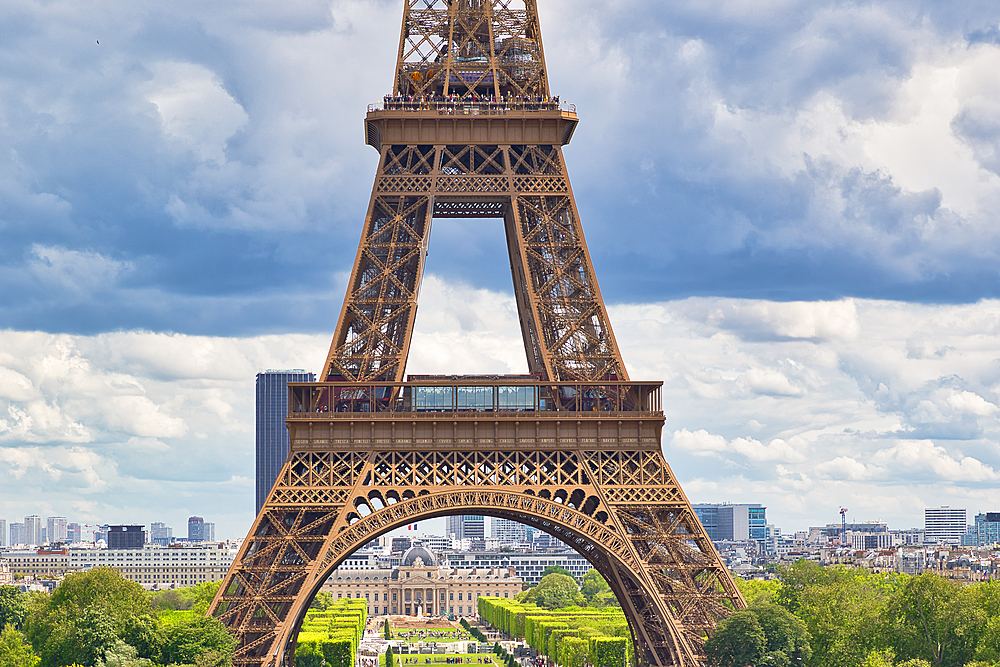 View of Eiffel Tower, foregrounded, rising above Champs de Mars and cityscape, against a partly cloudy sky.