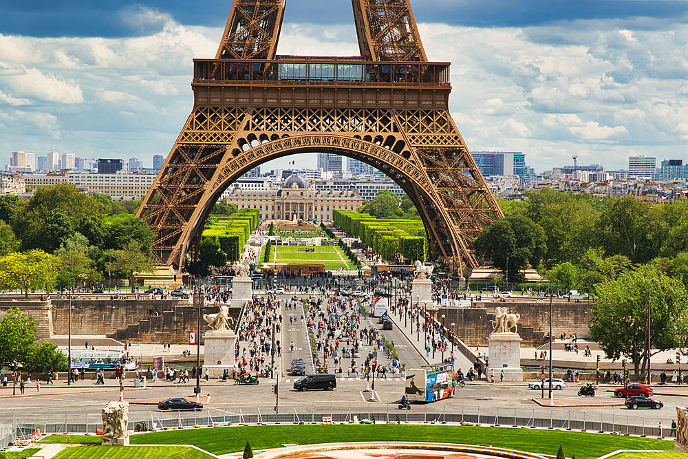 View through the Eiffel Tower archway shows a cityscape with a garden, buildings, many people, and traffic on a cloudy day.