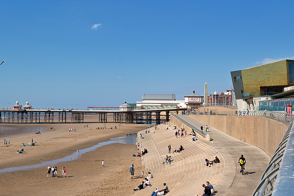 Beach scene with pier, modern building, and many people on a sunny day under a blue sky.