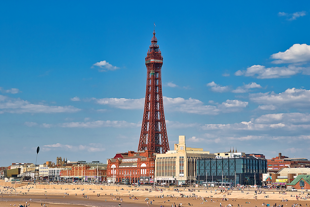 Wide shot: Blackpool Tower dominates the skyline, with buildings and a crowded beach below, under a blue sky with scattered clouds.