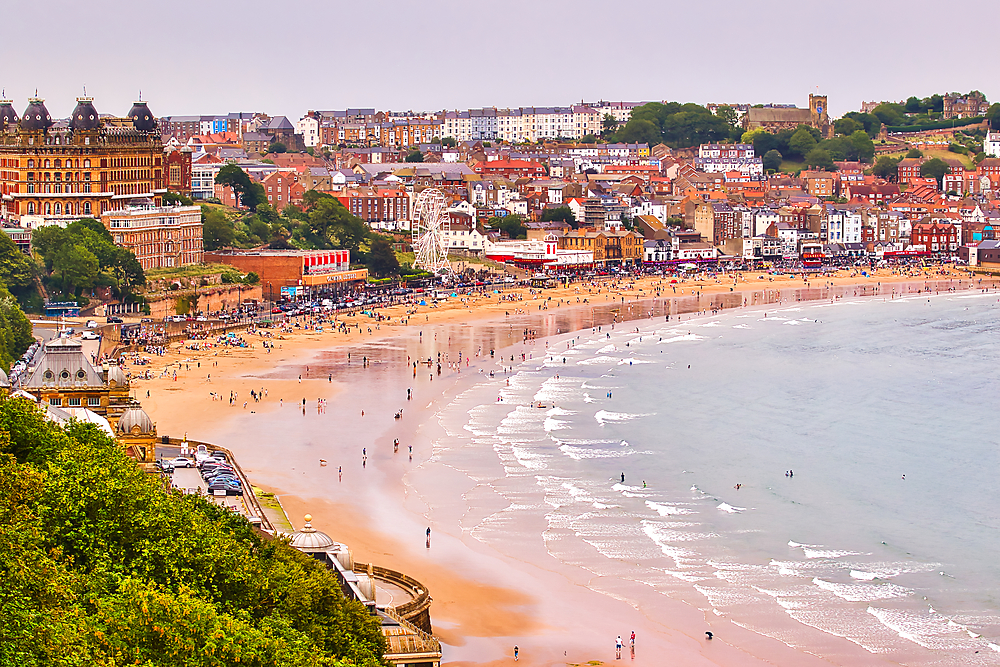 A panoramic view of a crowded beach with a town and a ferris wheel overlooking the water in Scarborough, UK.