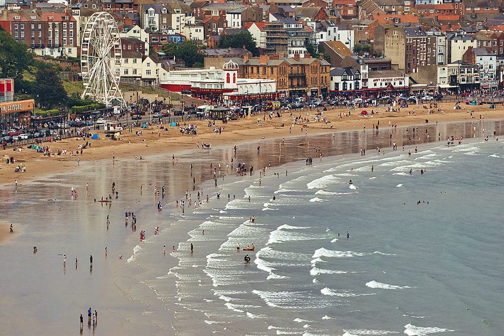 A bustling beach scene with many people, buildings, a Ferris wheel, and the ocean with waves in Scarborough, UK.