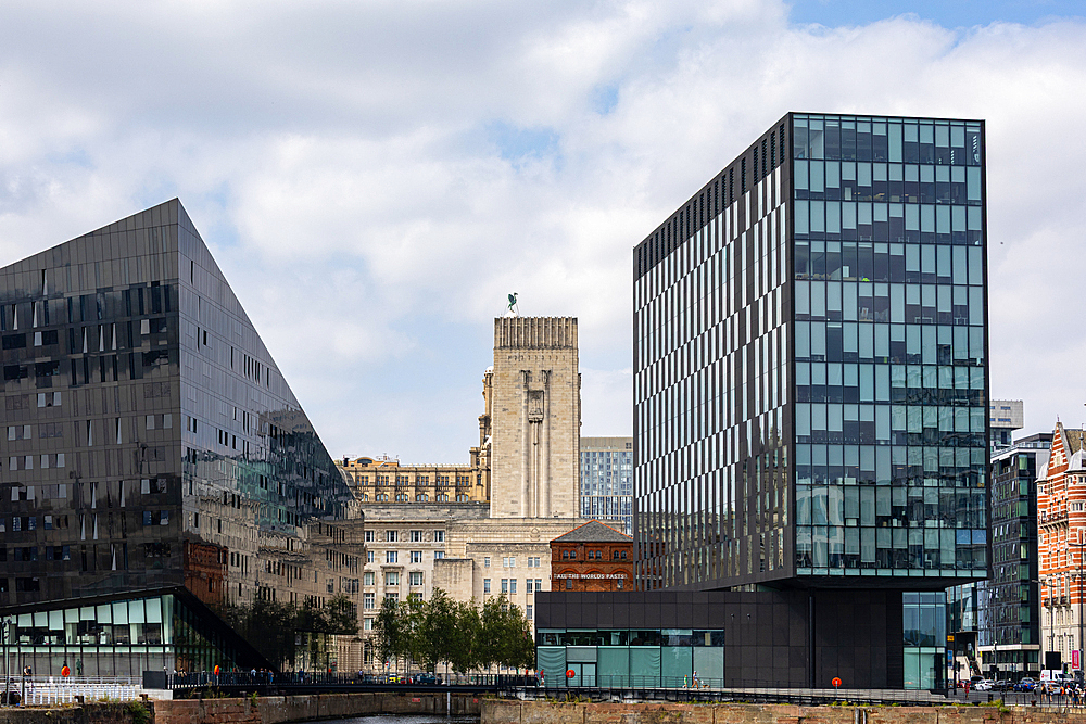 Modern glass buildings, a historic tower, and older architecture stand against a cloudy sky, reflected in dark water in Liverpool, UK.