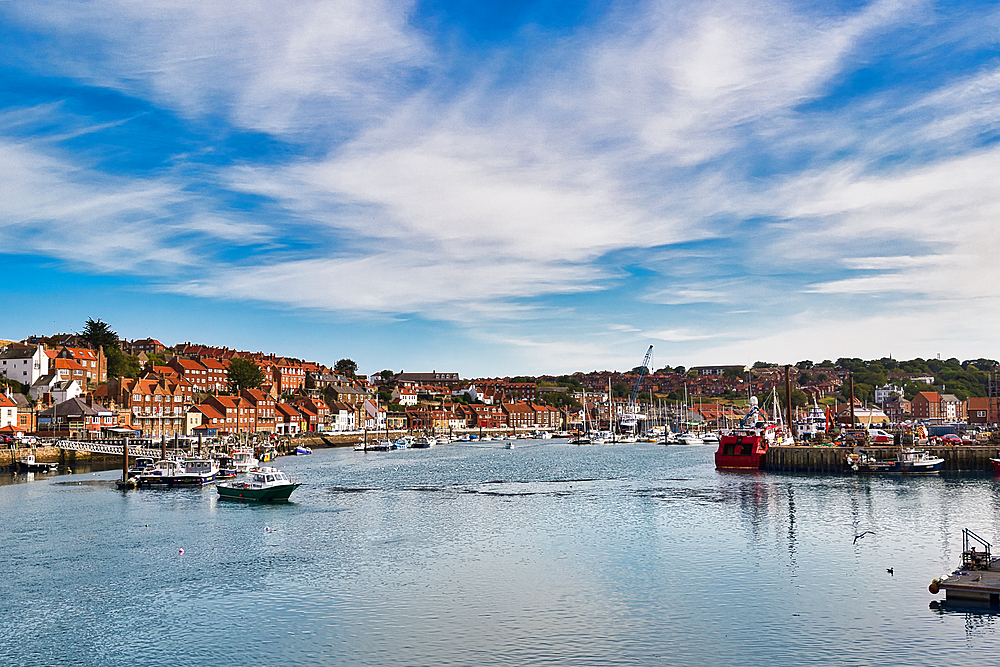 A picturesque harbor scene with a blue sky, white clouds, and a town with red-roofed buildings. Boats of various sizes are docked and in the water in Whitby, UK.