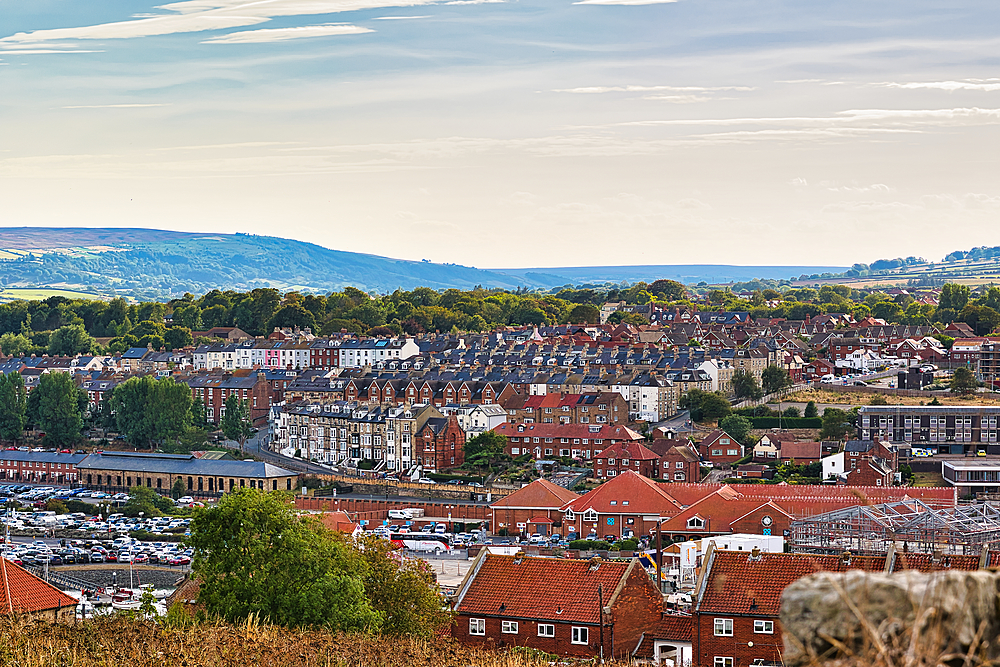 An aerial view of a coastal town featuring rows of houses with varied architectural styles and a backdrop of rolling green hills under a pale sky in Whitby, UK.