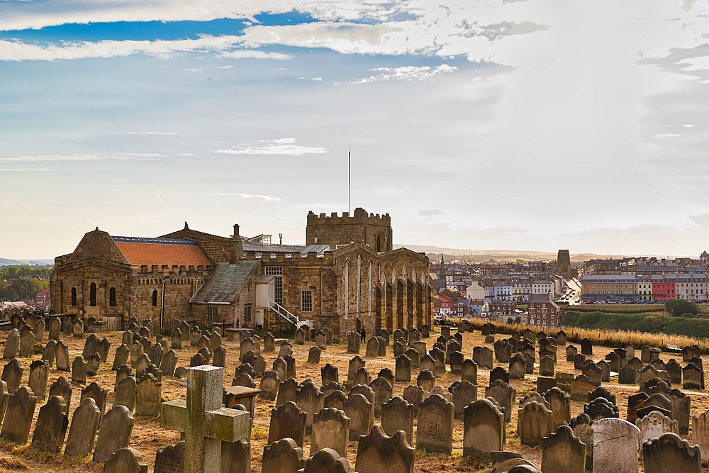 A stone church and a vast graveyard with weathered headstones overlook a colorful coastal town under a wide sky in Whitby, UK.