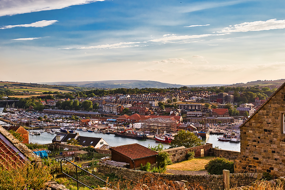 An elevated view captures a coastal town with a harbor filled with boats, nestled against rolling hills under a bright, partly cloudy sky in Whitby, UK.