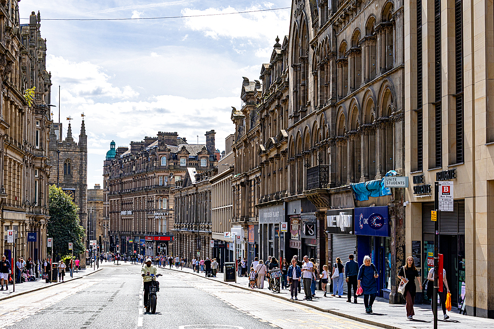 A wide street lined with historic stone buildings, featuring architectural details, shops, pedestrians, and a solitary cyclist in Newcastle upon Tyne, UK.