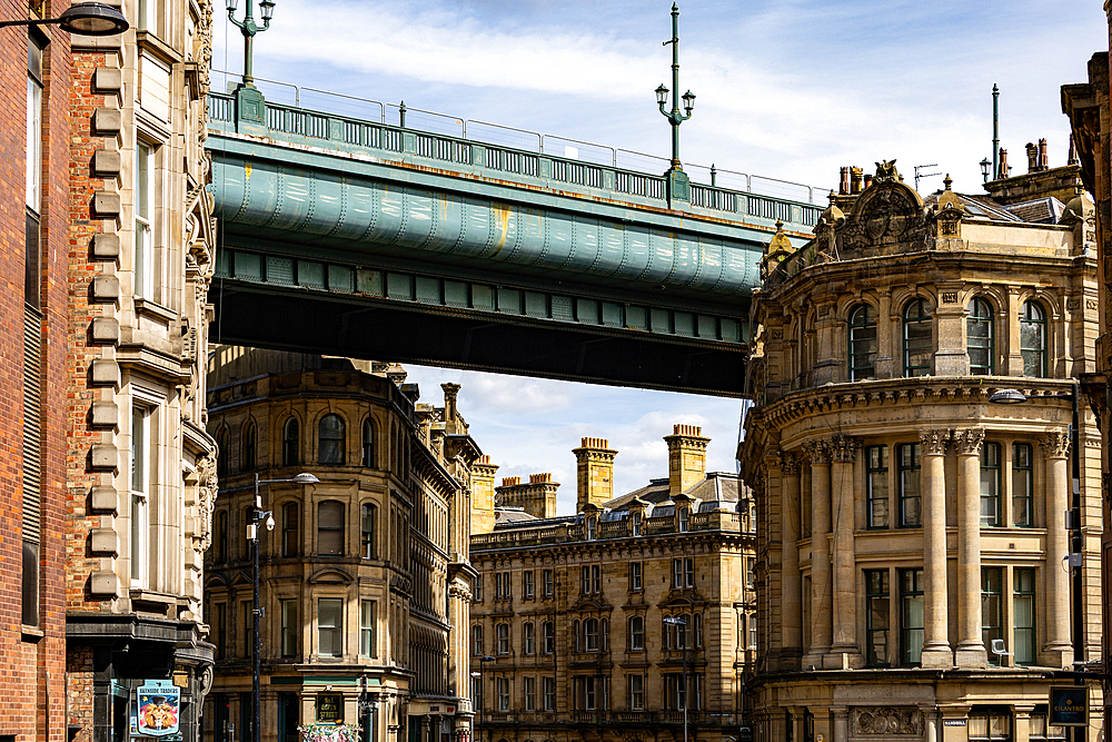 An urban scene featuring a large, teal-colored bridge spanning over ornate, stone buildings under a partly cloudy sky in Newcastle upon Tyne, UK.