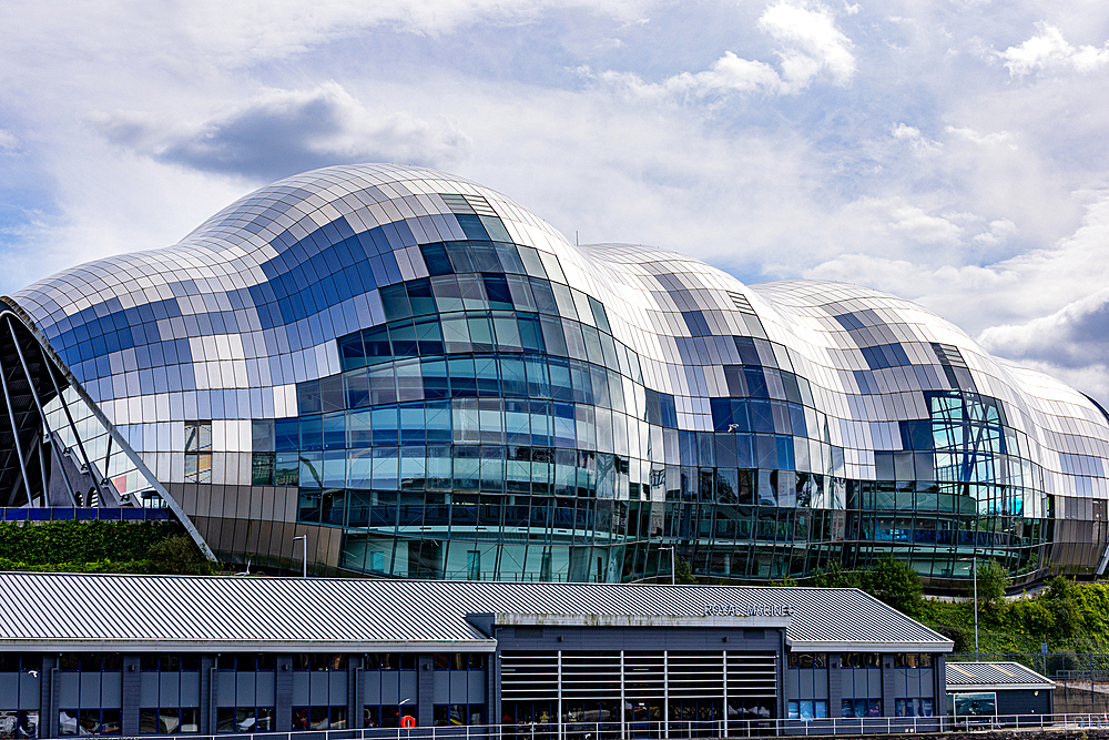 A modern, curved building with a distinctive tiled facade of silver, white, and blue panels, reflecting the cloudy sky in Newcastle upon Tyne, UK.