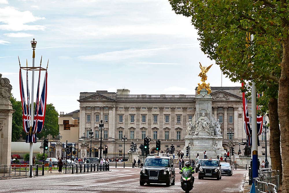Buckingham Palace with the Victoria Memorial and Union Jack flags, on a bright day with traffic and pedestrians on the street in London, UK.