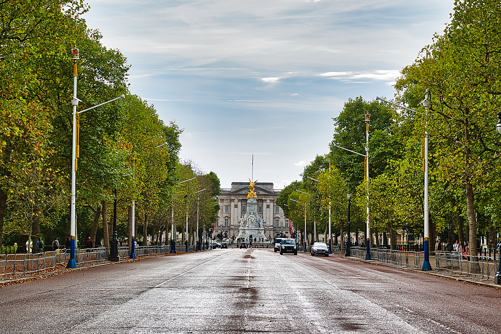 A wide, tree-lined avenue leads to a grand palace with a prominent golden statue at its center. The sky is overcast, and a few cars are visible on the wet road in London, UK.
