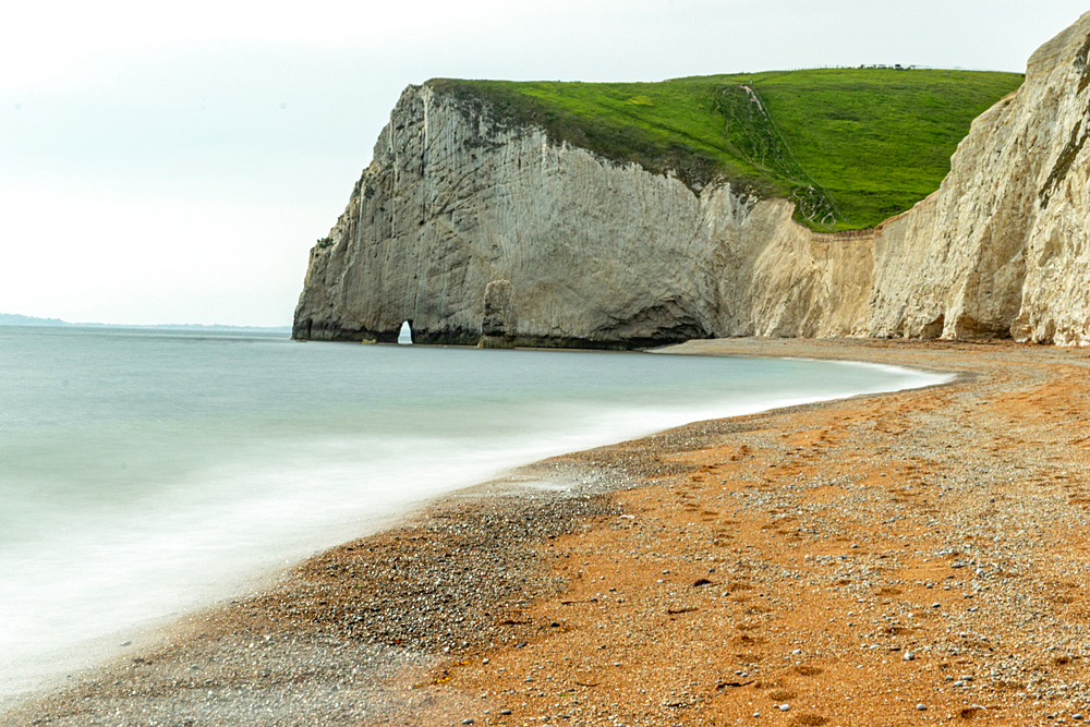 Serene beach with smooth waters, pebbles, and a towering cliff under a cloudy sky at Durdle Door, Jurassic Coast, UNESCO World Heritage Site, Dorset, England, United Kingdom, Europe