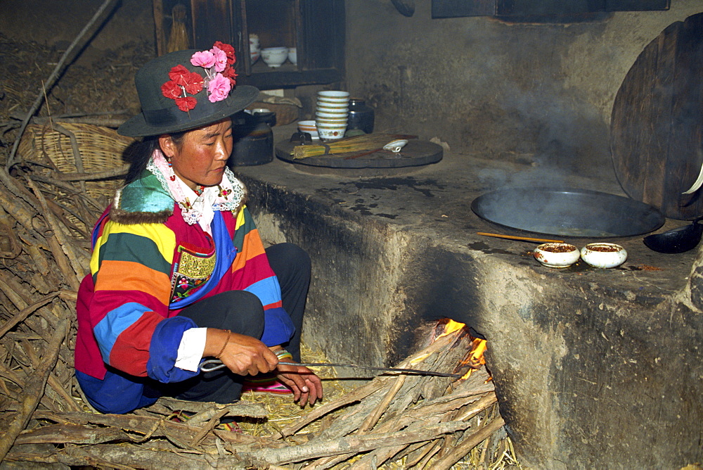 A Tu woman in bright clothes and hat stoking a wood fire for cooking in a kitchen near Xining, in Qinghai, China, Asia