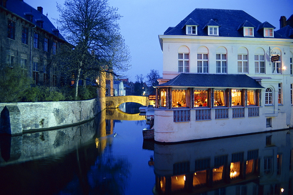 Reflections in the canals of restaurant and bridge, illuminated in the evening, in Bruges, Belgium, Europe