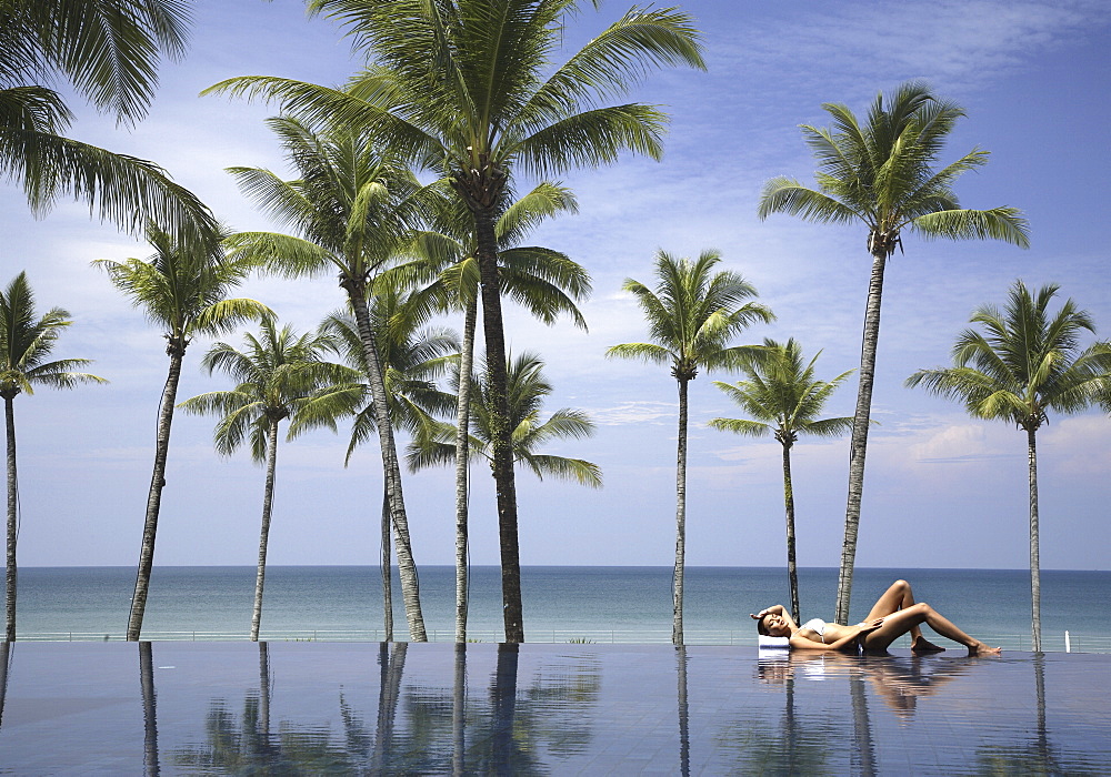 Young woman relaxing by pool with palm trees and the sea beyond
 