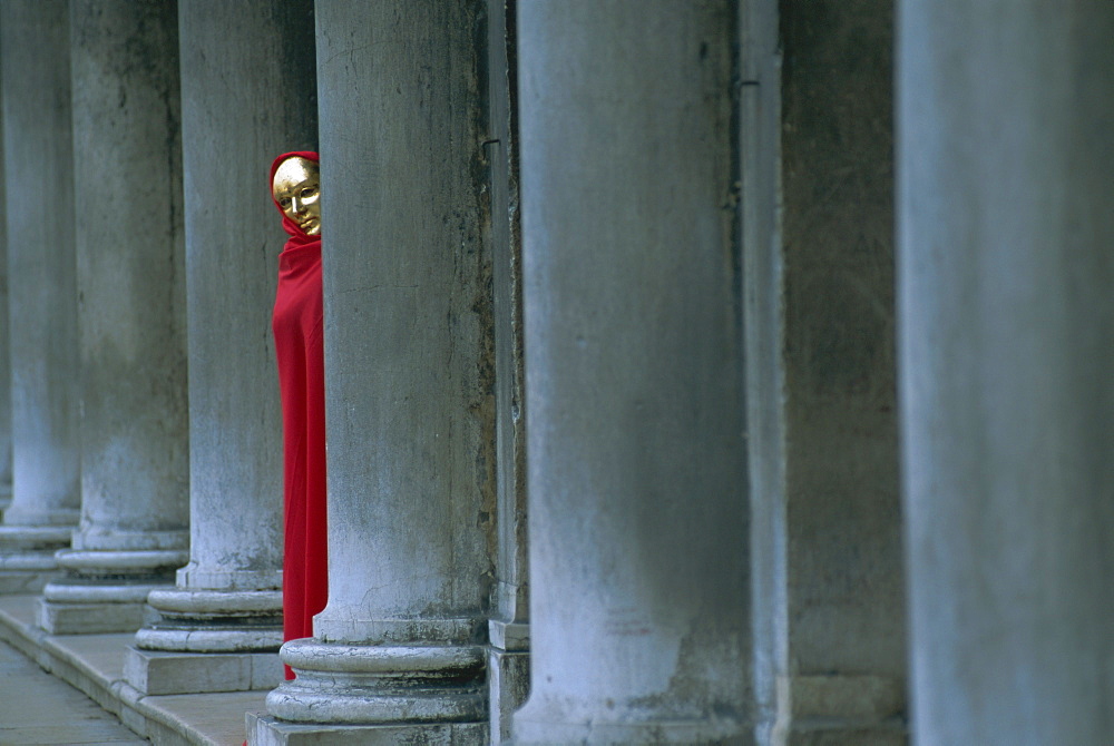 Carnival model in red cape and gold mask peering from columns in St. Mark's Square, Venice, UNESCO World Heritage Site, Veneto, Italy, Europe