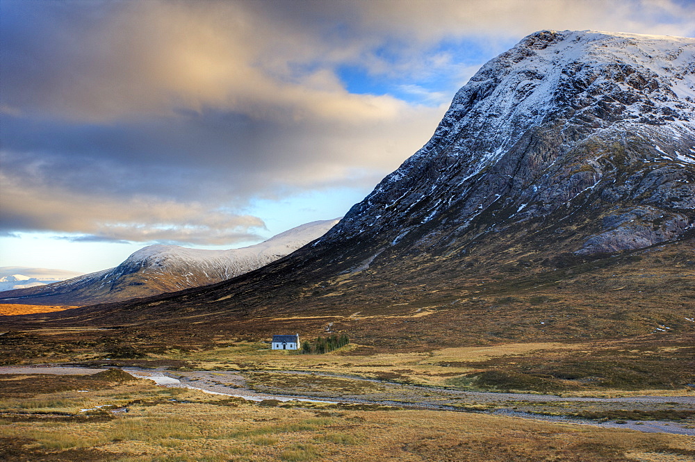 Winter view of Rannoch Moor showing lone whitewashed cottage on the bank of a river, dwarfed by snow-covered mountains, Rannoch Moor, near Fort William, Highland, Scotland, United Kingdom, Europe