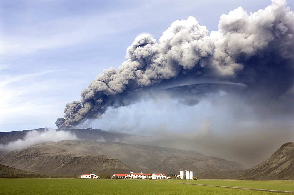 Farm buildings and green fields with the ash plume of the Eyjafjallajokull eruption in the distance, near Hella, southern area, Iceland, Polar Regions