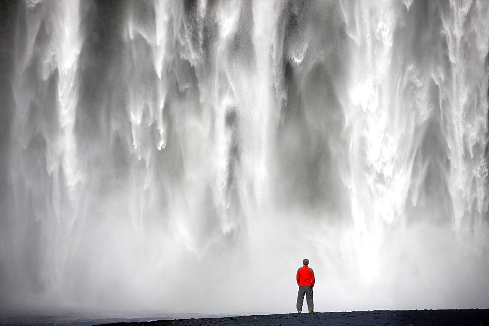 Man in red jacket standing in front of the 62m high Skogafoss waterfall near the village of Skogar, southern area, Iceland, Polar Regions