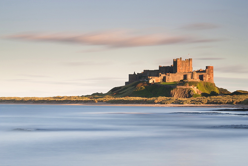 Bamburgh Castle bathed in golden evening light overlooking Bamburgh Bay with the sea filling the foreground, Northumberland, England, United Kingdom, Europe