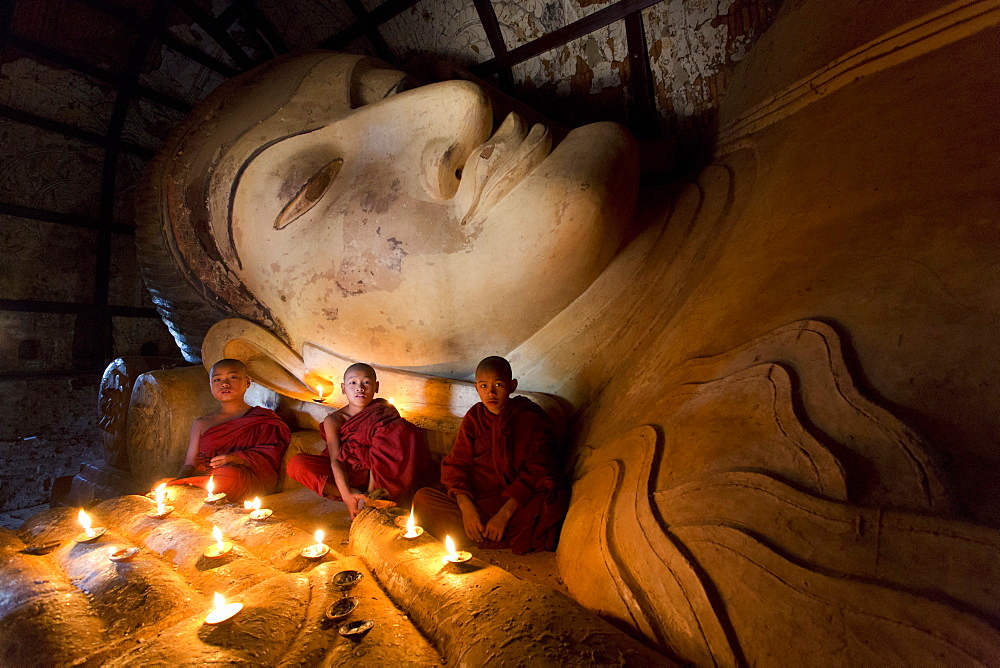 Three novice Buddhist monks sitting by a large reclining Buddha in the light of candles, near Shwesandaw Paya, Bagan, Myanmar (Burma), Asia