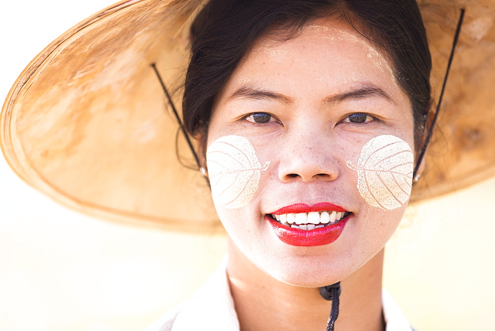 Backlit portrait of local woman wearing traditional bamboo hat and Thanaka face painting in the shape of leaves on her cheeks, near Mandalay, Myanmar (Burnma), Asia