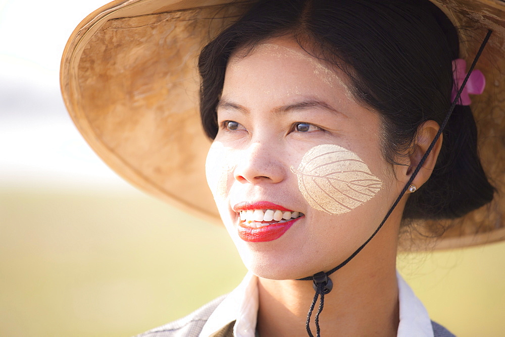 Backlit portrait of local woman wearing traditional bamboo hat and Thanaka face painting in the shape of leaves on her cheeks, near Mandalay, Myanmar (Burnma), Asia