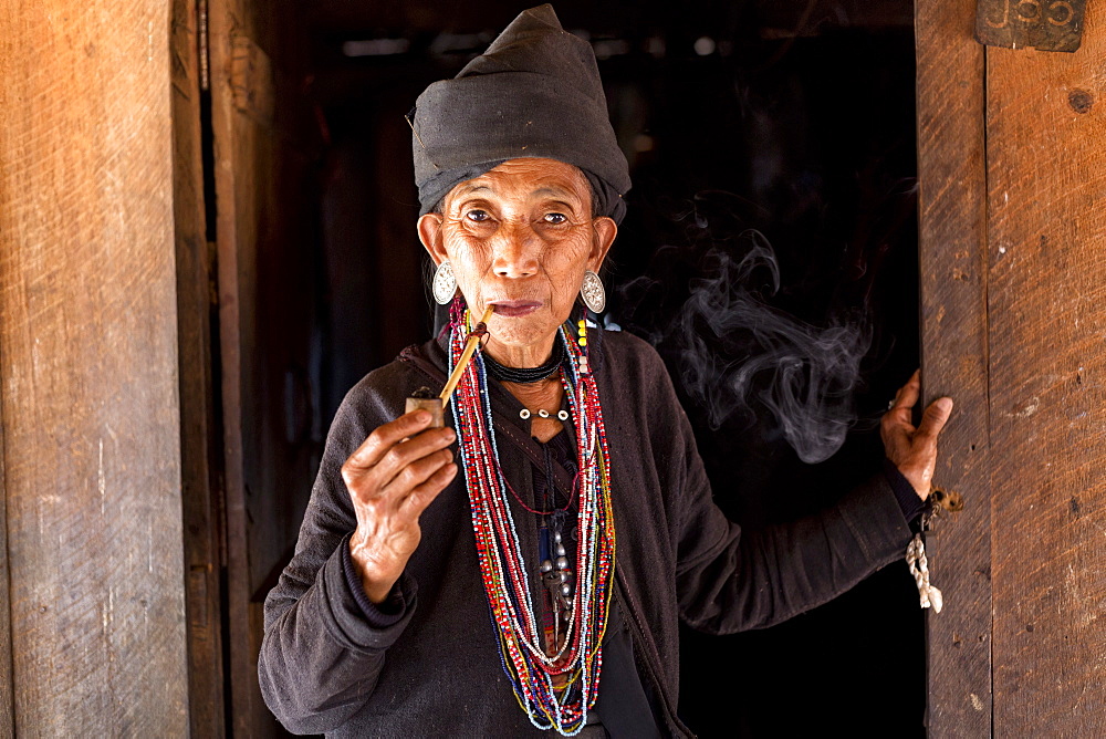 Woman of the Ann tribe in traditional black dress and colourful beads smoking a pipe in the doorway of her home in a hill village near Kengtung (Kyaingtong), Shan State, Myanmar (Burma), Asia