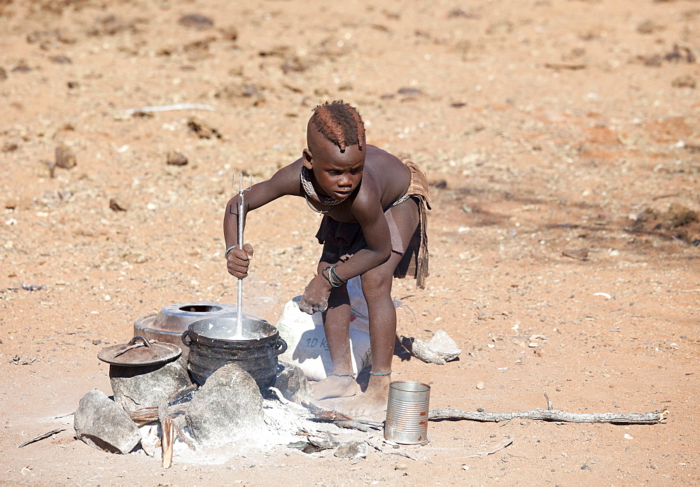 Young Himba boy cooking food on open fire in his village, Kunene Region (formerly Kaokoland) in the far north of Namibia, Africa