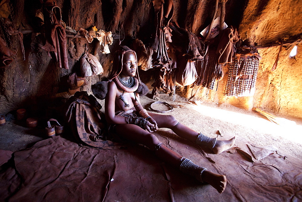 Young Himba woman inside a traditional mud dwelling hut wearing traditional dress and jewellery and with her skin covered in Otjize, a mixture of butterfat and ochre, Kunene Region, formerly Kaokoland, Namibia, Africa