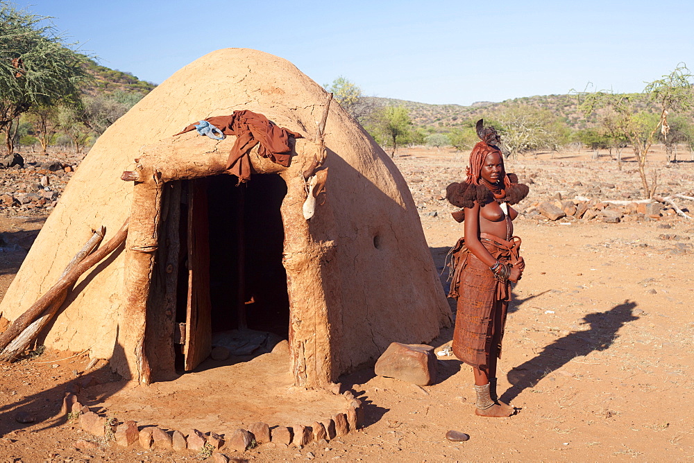 Young Himba woman standing outside her traditional mud-covered dwelling in a Himba village in the Kunene Region (formerly Kaokoland) in the far north of Namibia